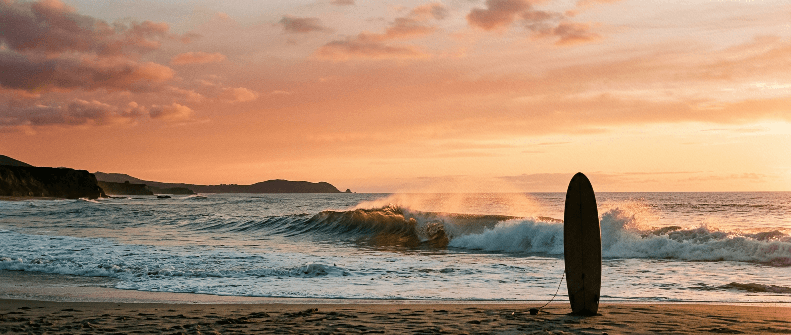 A surfboard planted in the sand as a wave breaks at sunset